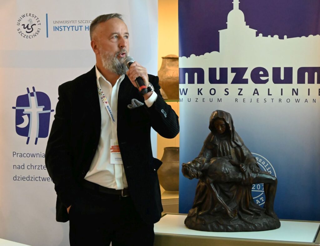 Prelegent przemawia do publiczności, trzymając mikrofon, na tle ekspozycji muzealnej (kliknięcie powiększy zdjęcie).