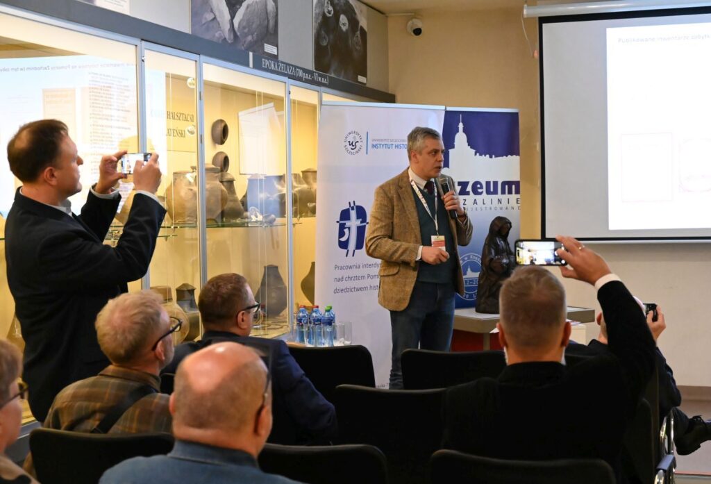 Prelegent przemawia do uczestników konferencji w sali muzealnej, część osób robi zdjęcia telefonami (kliknięcie powiększy zdjęcie).