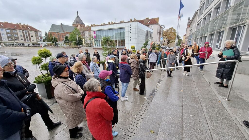 Grupa uczestników spaceru historycznego stoi na schodach przed Ratuszem podczas opowieści przewodnika (kliknięcie powiększy zdjęcie).