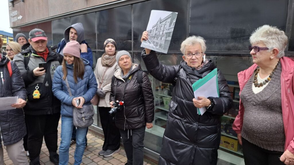 Uczestnicy spaceru historycznego oglądają archiwalne zdjęcie prezentowane przez przewodniczkę (kliknięcie powiększy zdjęcie).