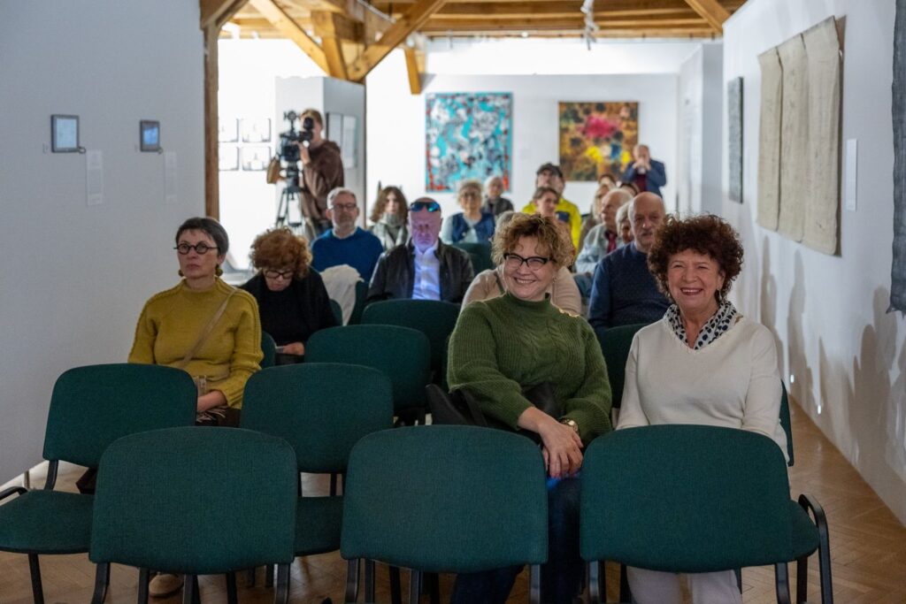 Publiczność siedzi na zielonych krzesłach w sali wystawowej. Kilka osób uśmiecha się do obiektywu. W tle stoi operator kamery oraz widać prace dostępne na wystawie (kliknięcie spowoduje powiększenie zdjęcia)