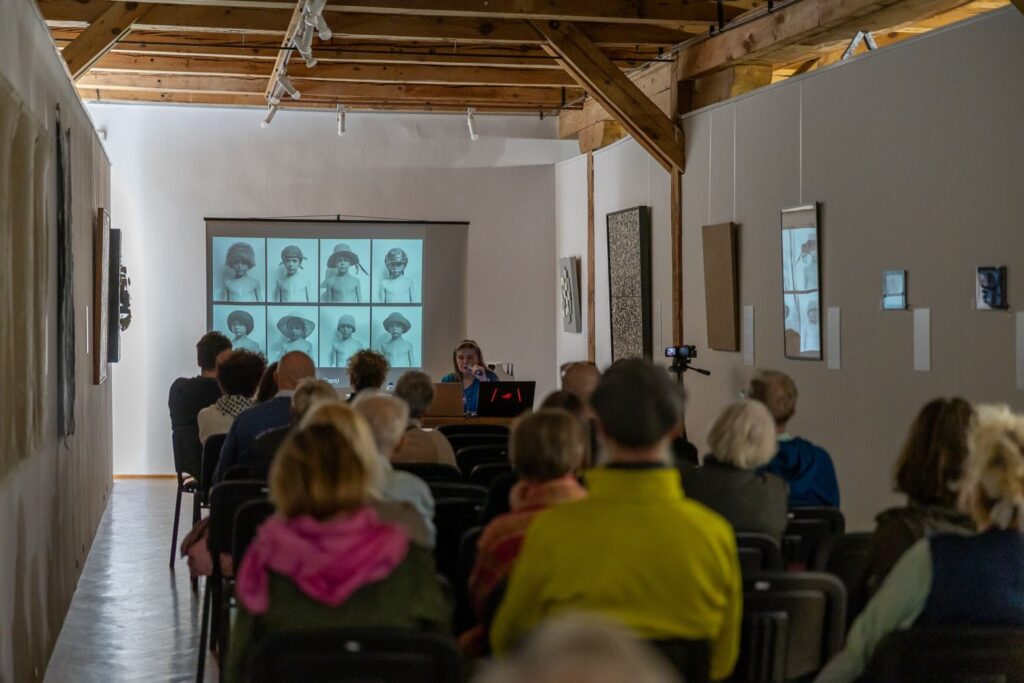 Widok z tyłu sali podczas prelekcji, na sali siedzi widownia. Na ekranie widać czarno-białe fotografie i projekcję (kliknięcie spowoduje powiększenie zdjęcia)