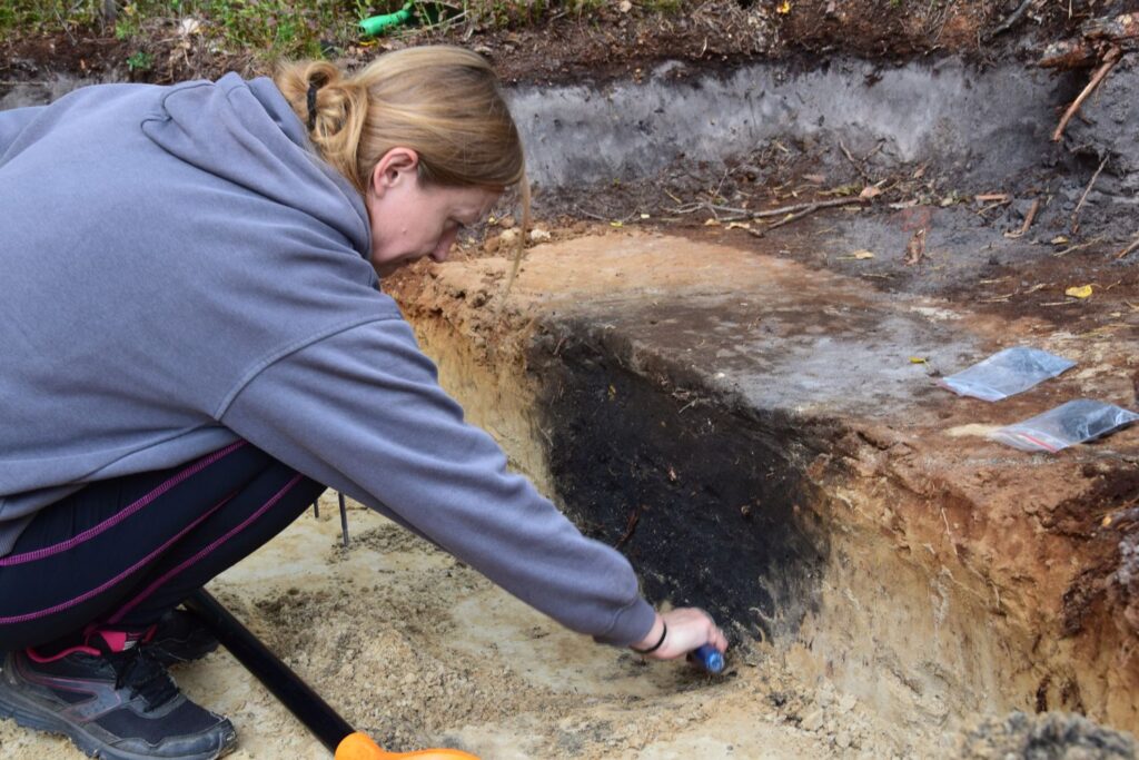 Kobieta pracuje w wykopie archeologicznym i jakimś narzędziem odsłania ciemniejszą warstwę ziemi (kliknięcie spowoduje powiększenie fotografii).
