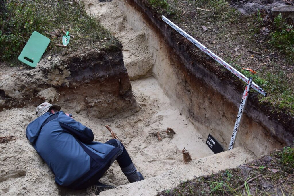 Mężczyzna leży w wykopie archeologicznym i fotografuje odnalezione artefakty. W wykopie znajdują się linie pomiarowe (kliknięcie spowoduje powiększenie fotografii).