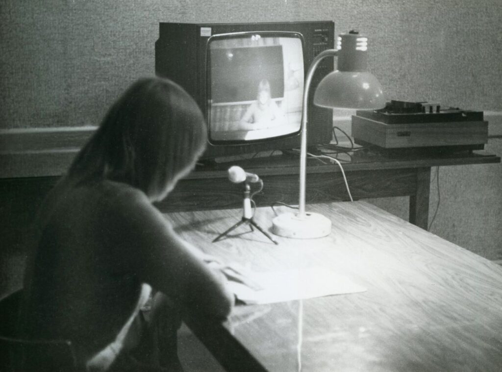 Kobieta siedzi przy biurku i czyta do mikrofonu, w tle na stole stoi telewizor, na którym wyświetlany jest obraz (kliknięcie powiększy zdjęcie).