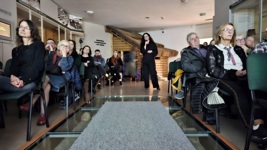 Uczestnicy wydarzenia siedzą po obu stronach przeszklonej podłogi w sali muzealnej i słuchają prowadzącej wykład o Halloween (kliknięcie powiększy zdjęcie)