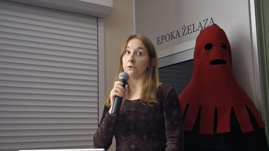 Kobieta trzymająca mikrofon przemawia do publiczności, za nią stoi czerwona dekoracja przypominająca ducha, na ścianie widoczny napis „Epoka żelaza” (kliknięcie powiększy zdjęcie).