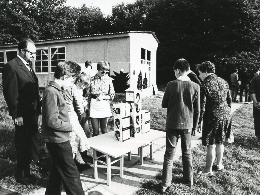 Czarno-biała fotografia przedstawiająca grupę osób oglądających prace ustawione na stolikach na świeżym powietrzu. W tle widoczny jest pawilon wystawowy oraz drzewa, a część uczestników stoi wokół ekspozycji złożonej z niewielkich obiektów i fotografii (kliknięcie powiększy zdjęcie).