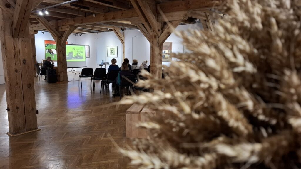 Sala wystawowa - widok ogólny. W tle siedzą osoby i słuchają wykładu. Na pierwszym planie, po prawej, fragment wieńca dożynkowego (kliknięcie spowoduje powiększenie zdjęcia)
