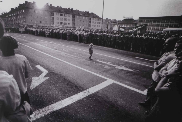 Czarno-biała fotografia przedstawiająca małe dziecko stojące samotnie na środku szerokiej ulicy, po której obu stronach stoją tłumy ludzi obserwujących wydarzenie lub zgromadzenie (kliknięcie powiększy zdjęcie).