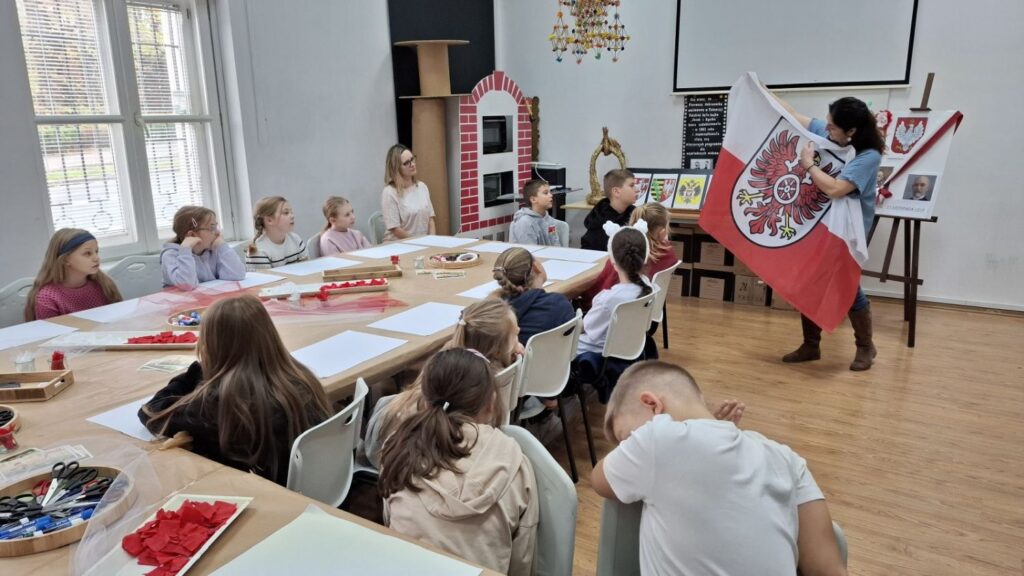 Dzieci siedzą przy stołach podczas warsztatów, a prowadząca pokazuje biało-czerwoną flagę z orłem i opowiada o jej symbolice (kliknięcie powiększy zdjęcie).