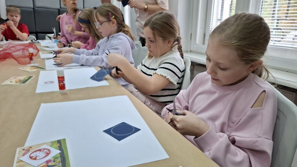 Dziewczynki siedzą przy stole i wycinają z papieru elementy potrzebne do wykonania biało-czerwonej kokardy narodowej (kliknięcie powiększy zdjęcie).