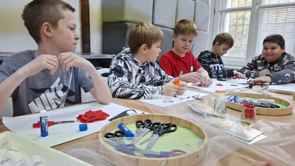 Chłopcy pracują przy stole z materiałami plastycznymi, wycinając elementy do przygotowania kokard narodowych (kliknięcie powiększy zdjęcie).
