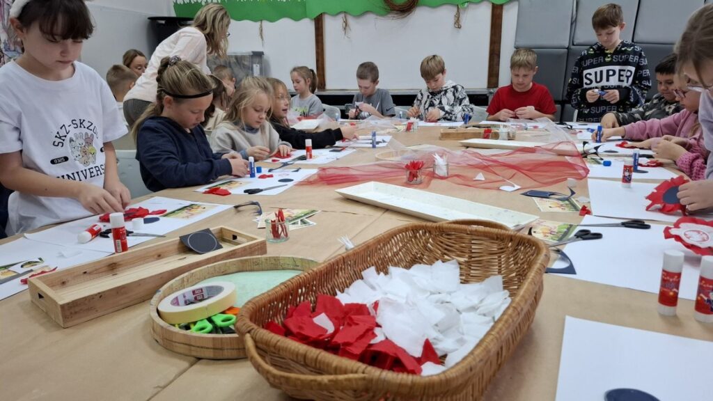 Grupa dzieci skupiona przy dużym stole podczas wspólnego wykonywania biało-czerwonych ozdób z papieru i wstążek (kliknięcie powiększy zdjęcie).