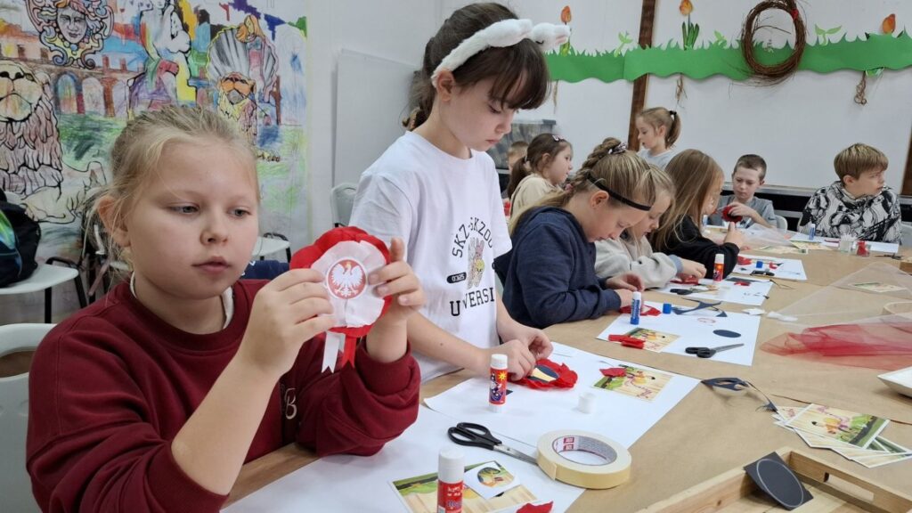 Dziewczynka pokazuje gotową biało-czerwoną kokardę narodową, a wokół niej inne dzieci pracują nad swoimi pracami plastycznymi (kliknięcie powiększy zdjęcie).