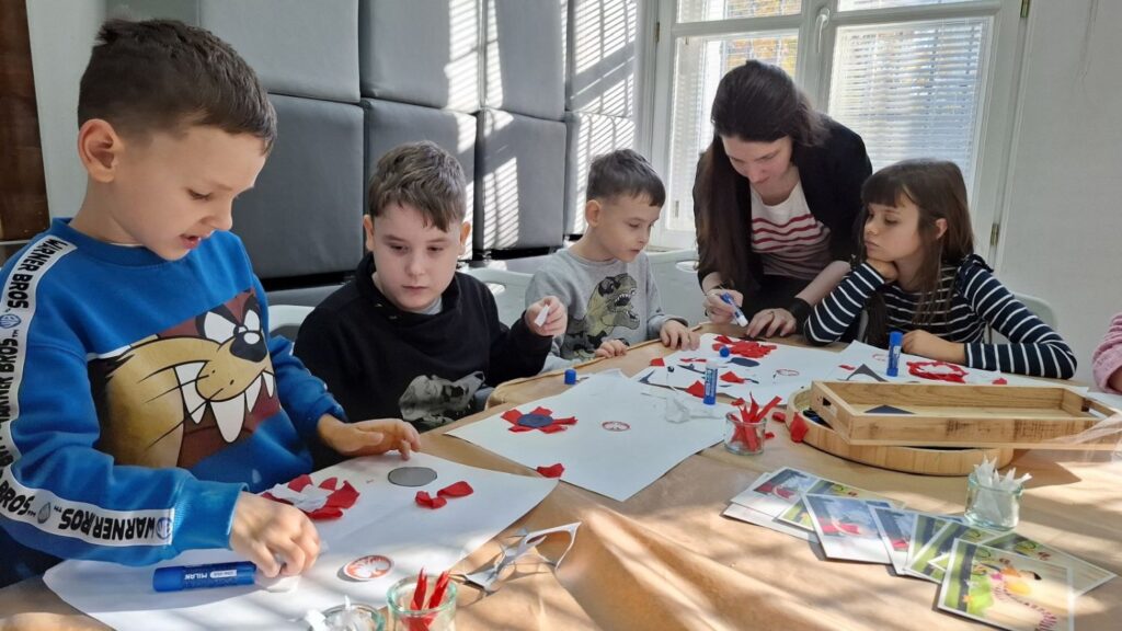 Grupa dzieci wraz z prowadzącą pracuje nad swoimi pracami plastycznymi, przygotowując biało-czerwone ozdoby (kliknięcie powiększy zdjęcie).