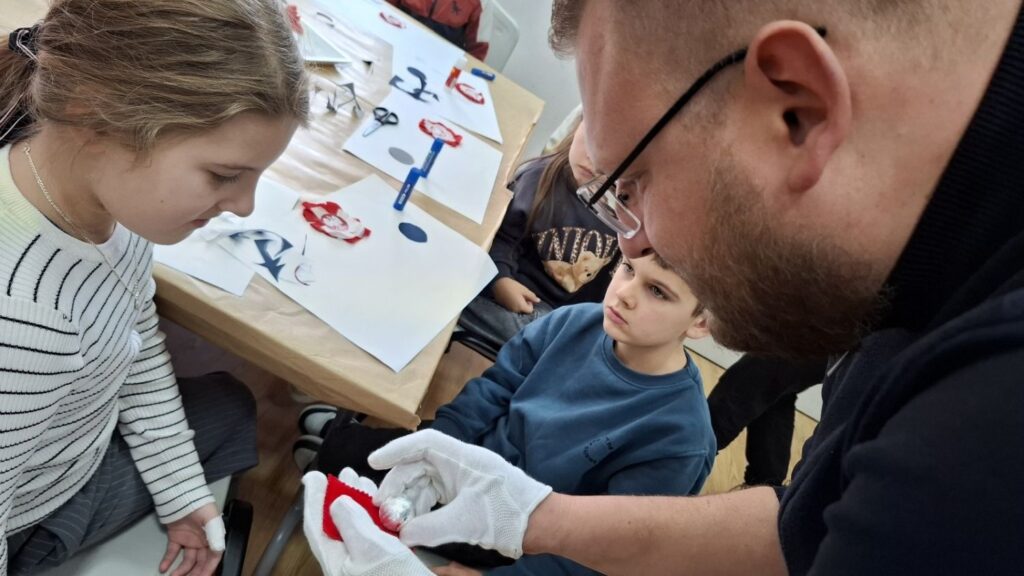 Kierownik Działu Historii w rękawiczkach pokazuje dzieciom element kokardy narodowej i tłumaczy sposób jej wykonania (kliknięcie powiększy zdjęcie).