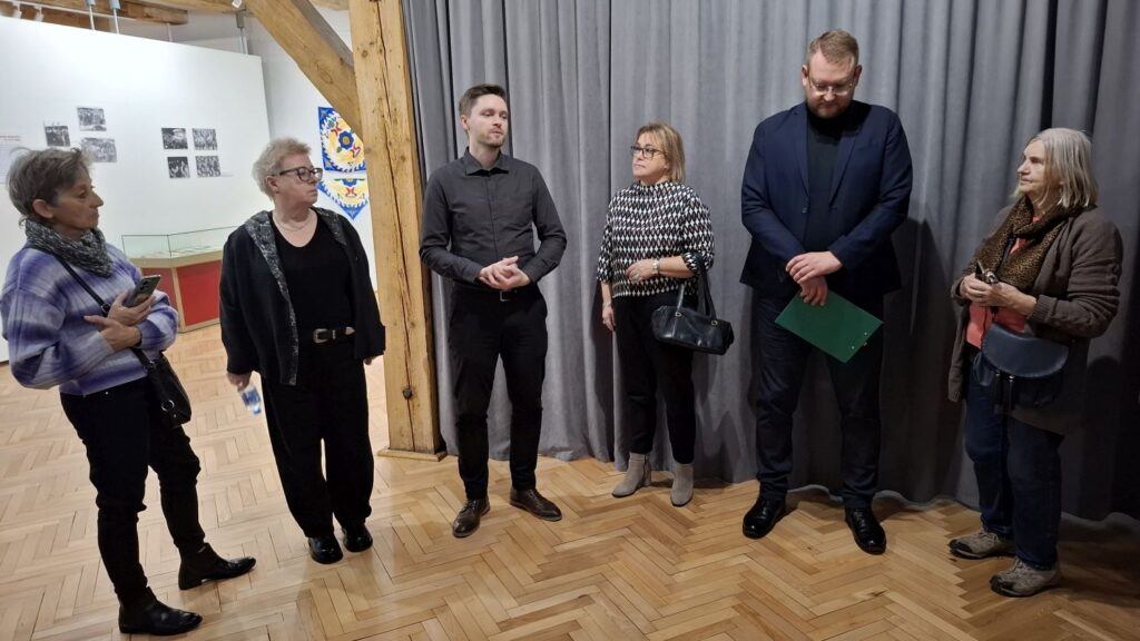 Osoby prowadzące oprowadzanie po wystawie stoją obok siebie, w sali muzealnej, i witają uczestników spotkania (kliknięcie powiększy zdjęcie).