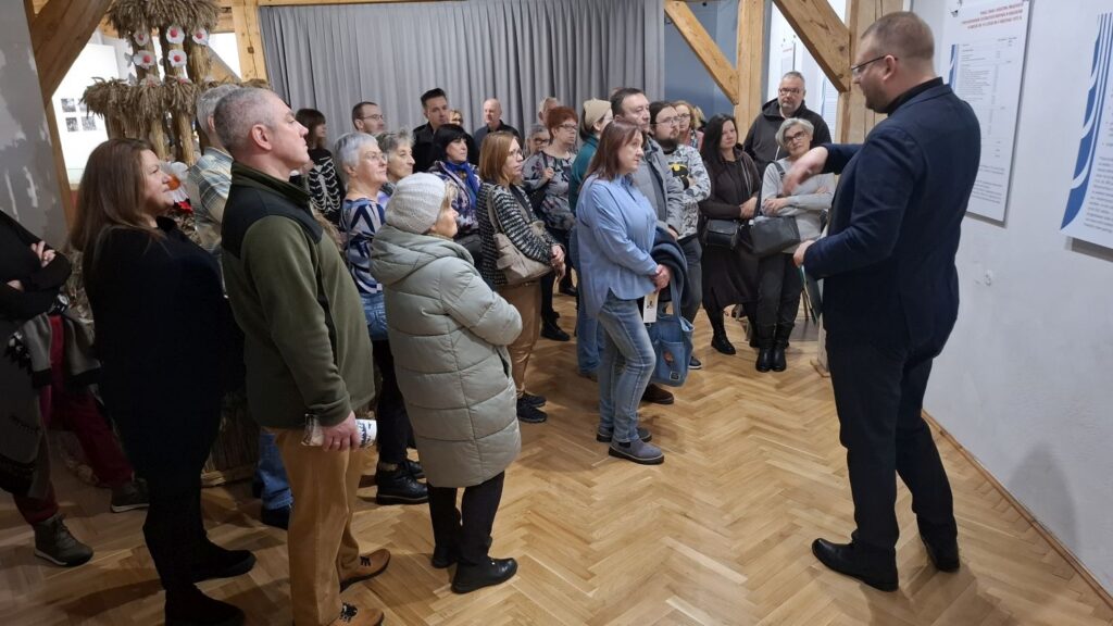 Kierownik działu historycznego opowiada o ekspozycji, a zgromadzeni uczestnicy słuchają jego wyjaśnień przy planszy z materiałami wystawowymi (kliknięcie powiększy zdjęcie).
