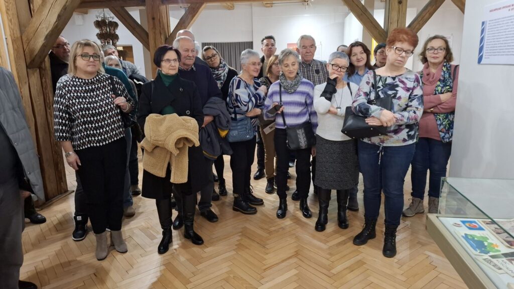 Zwiedzający stoją w sali muzealnej i słuchają opowieści przewodnika podczas oprowadzania po wystawie (kliknięcie powiększy zdjęcie).