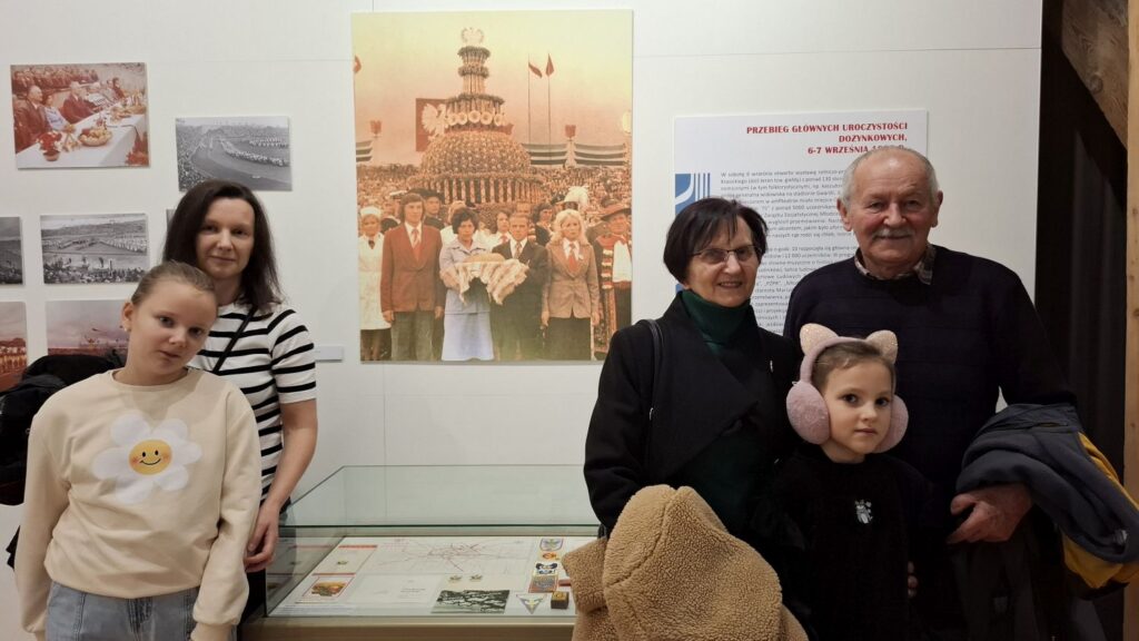 Rodzina Edwarda stoi przy gablocie z materiałami wystawowymi na tle planszy z archiwalną fotografią z Dożynek Centralnych 1975 (kliknięcie powiększy zdjęcie).