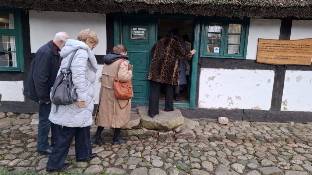 Uczestnicy spotkania wchodzą do zabytkowej chaty o bielonych ścianach i zielonych drzwiach podczas zwiedzania poświęconego tradycjom bożonarodzeniowym (kliknięcie powiększy zdjęcie).