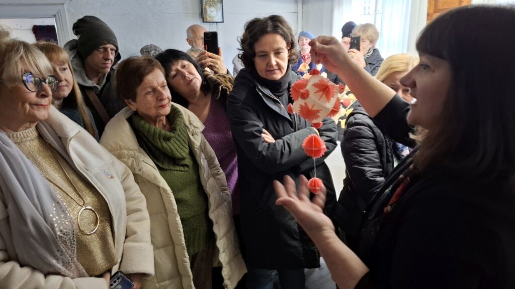 Przewodniczka pokazuje uczestnikom tradycyjną papierową ozdobę choinkową, a zgromadzone osoby przyglądają się jej z bliska (kliknięcie powiększy zdjęcie).