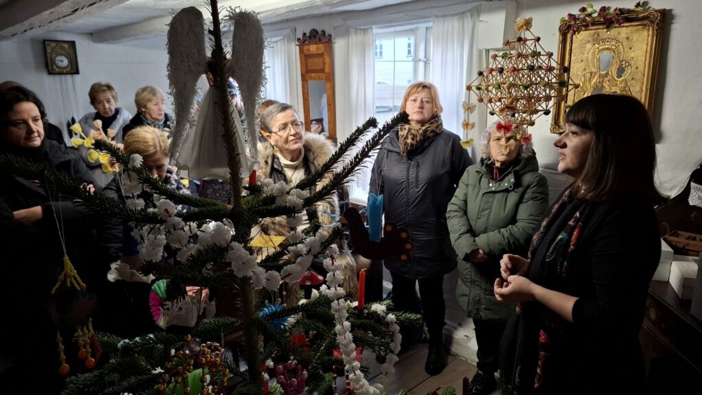 Uczestnicy stoją wokół dużej choinki ozdobionej tradycyjnymi dekoracjami, słuchając opowieści o dawnych zwyczajach bożonarodzeniowych (kliknięcie powiększy zdjęcie).
