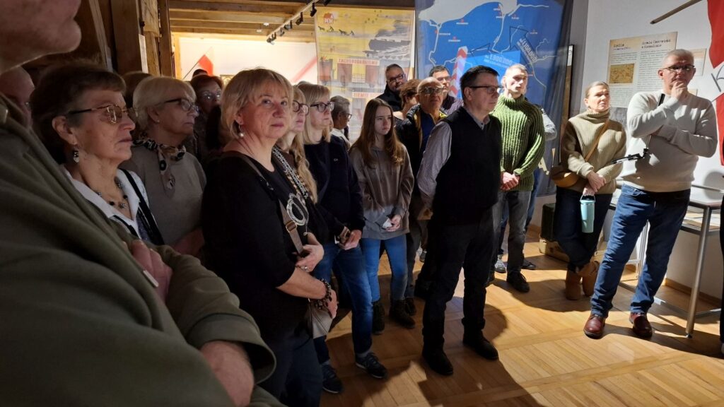Zgromadzeni uczestnicy wydarzenia słuchający oprowadzania w sali muzealnej; ludzie stoją w półokręgu wśród eksponatów (kliknięcie powiększy zdjęcie).