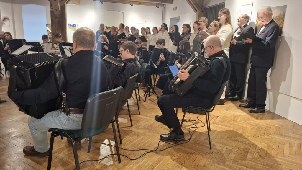 Muzycy akompaniujący chórowi, siedzący na krzesłach z instrumentami podczas koncertu w galerii (kliknięcie powiększy zdjęcie)