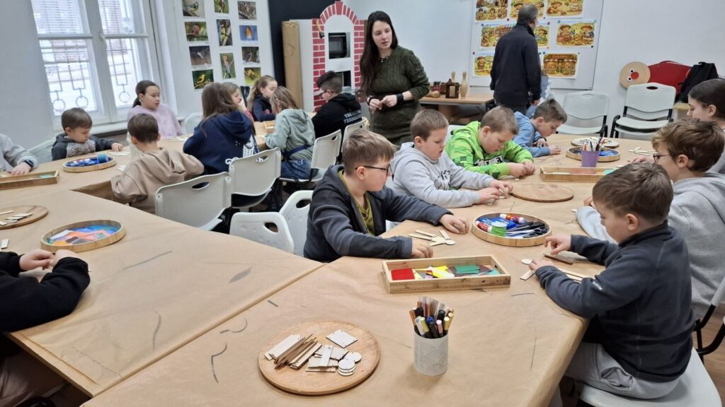 Sala warsztatowa z grupą dzieci pracujących przy dużych stołach nad drewnianymi zabawkami, w tle prowadząca nadzoruje zajęcia, a na blatach leżą farby, kredki i drewniane elementy (kliknięcie powiększy zdjęcie).