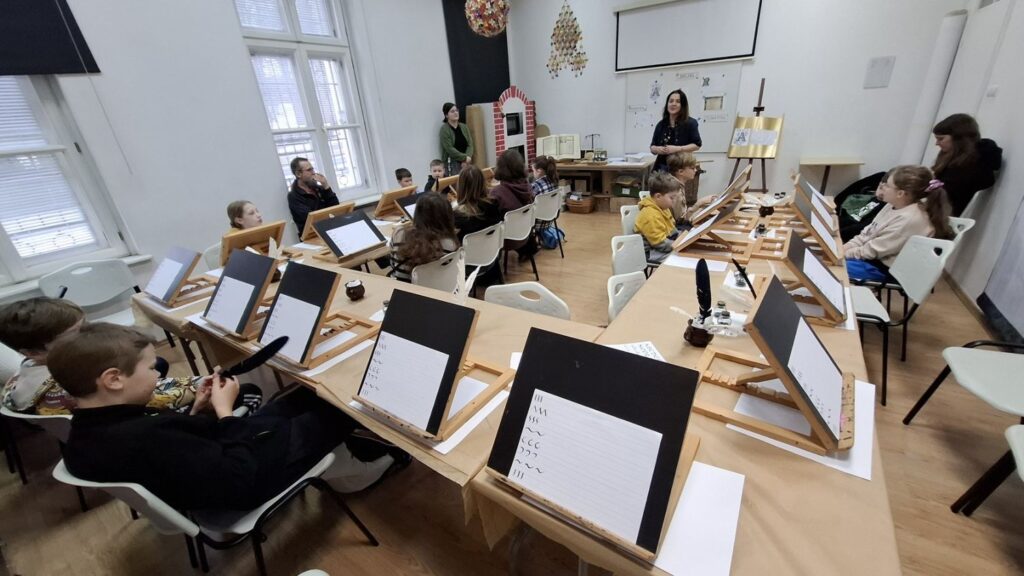 Dzieci siedzą przy sztalugach i ćwiczą kaligrafię, przepisując litery na kartkach podczas zajęć prowadzonych przez instruktorkę (kliknięcie powiększy zdjęcie).