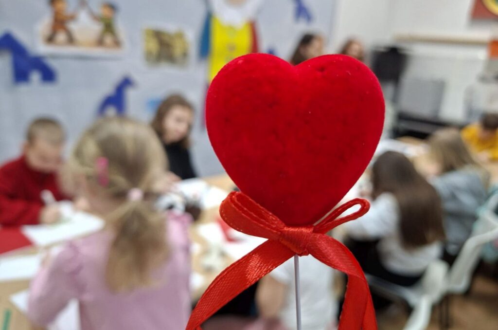 Czerwone serce na patyczku z kokardką na pierwszym planie, w tle dzieci pracujące nad walentynkowymi kartkami (kliknięcie powiększy zdjęcie).