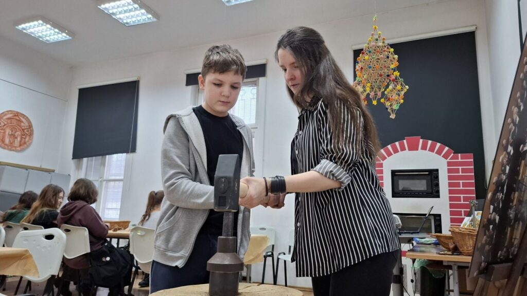 Chłopiec, z pomocą osoby prowadzącej zajęcia, wybija wzory na metalowym krążku (kliknięcie powiększy zdjęcie).