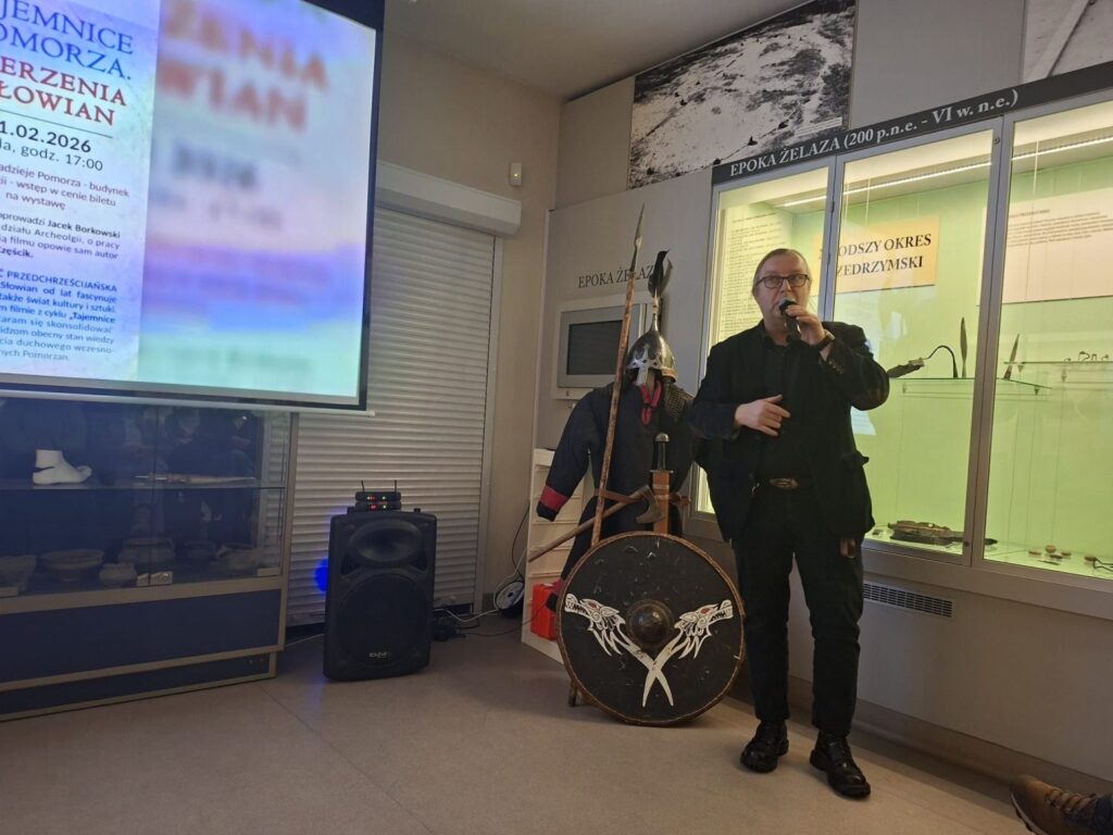 Prelegent trzymający mikrofon przemawia do publiczności w sali muzealnej, obok stoją elementy rekonstrukcji uzbrojenia oraz tarcza (kliknięcie powiększy zdjęcie).