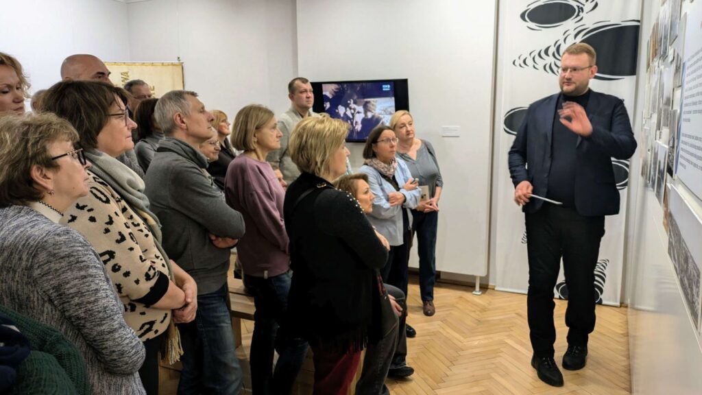 Przewodnik wskazuje na planszę z materiałami archiwalnymi, a zgromadzeni uczestnicy z uwagą śledzą jego wyjaśnienia (kliknięcie powiększy zdjęcie).