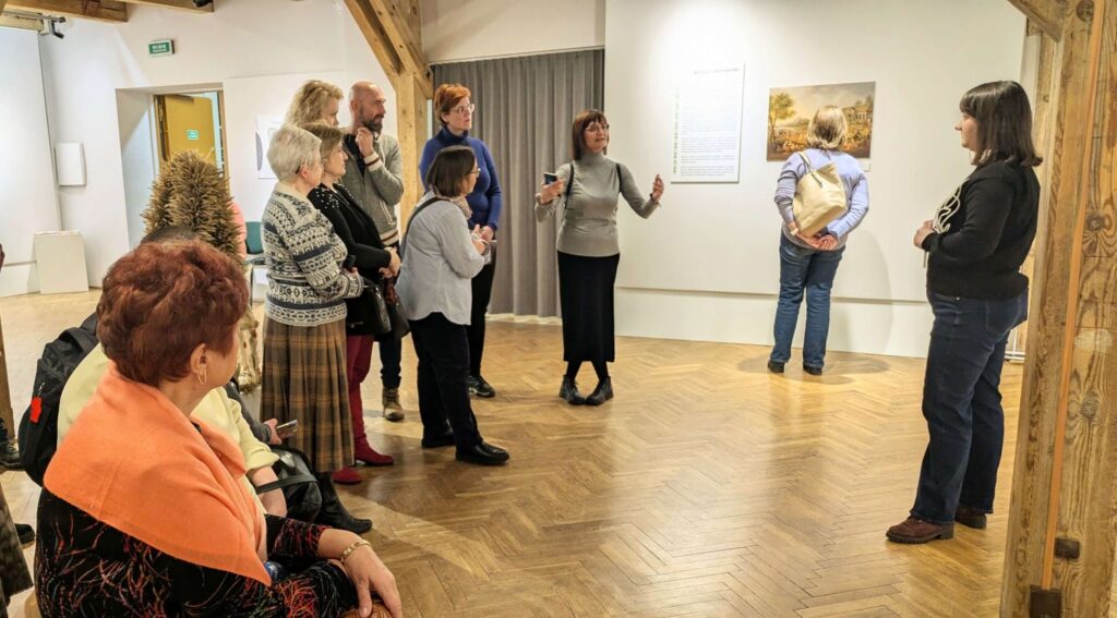 Osoba oprowadzająca po wystawie omawia prezentowane na niej prace, podczas gdy grupa ustawia się półkolem wokół niej (kliknięcie powiększy zdjęcie).