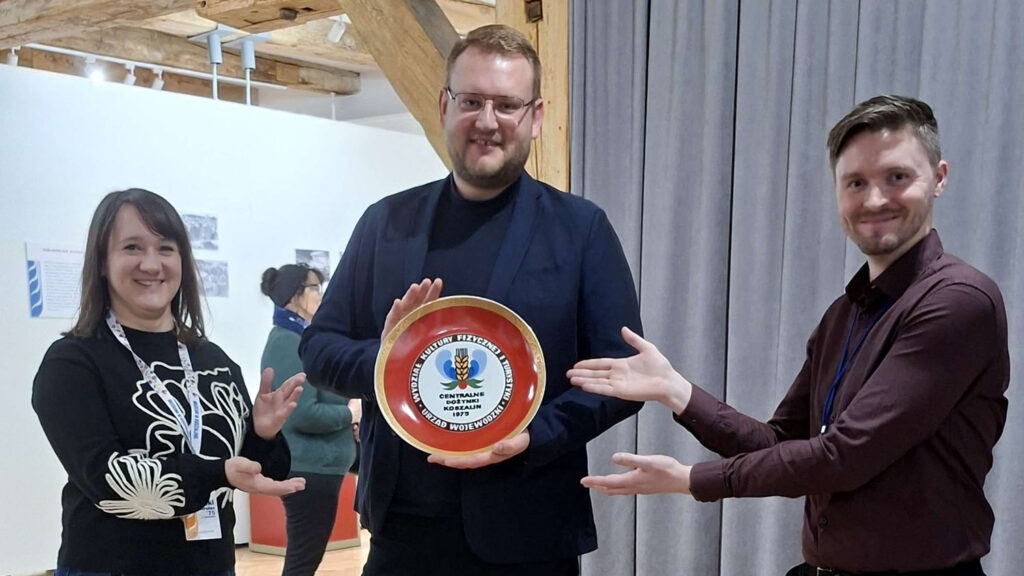 Trzy osoby stoją w sali wystawowej i prezentują artefakt przekazany do muzeum - okolicznościową pamiątkową tablicę z logo Centralnych Dożynek 1975 w Koszalinie, trzymając ją wspólnie i wskazując na nią dłońmi (kliknięcie powiększy zdjęcie).