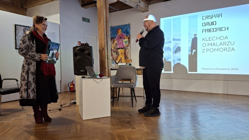 Prowadzący spotkanie oraz dyrektor Muzeum stoją przed publicznością w sali wystawowej, w tle widoczna prezentacja multimedialna poświęcona twórczości Caspara Davida Friedricha (kliknięcie powiększy zdjęcie).