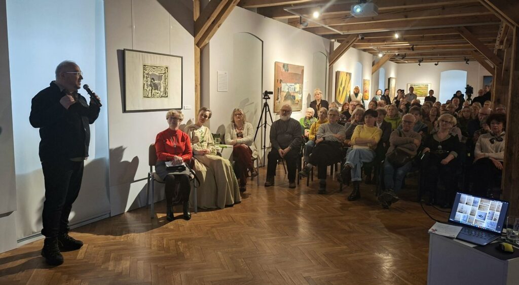 Prelegent przemawiający do licznie zgromadzonej publiczności w przestrzeni galerii, obok ścian z prezentowanymi pracami artystycznymi (kliknięcie powiększy zdjęcie).