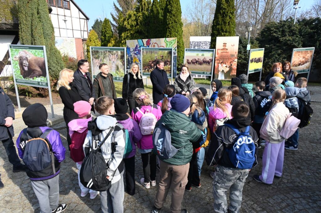 Grupa dzieci wraz z opiekunami stoi na dziedzińcu i słucha informacji o rozpoczęciu wydarzenia przez dyrektor muzeum, w tle wystawa fotograficzna (kliknięcie powiększy zdjęcie).