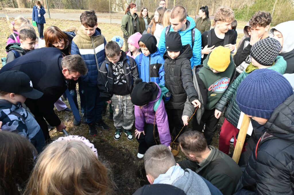 Dzieci z bliska przyglądają się sadzeniu drzewa, w tle stoją pozostali uczestnicy wydarzenia (kliknięcie powiększy zdjęcie).