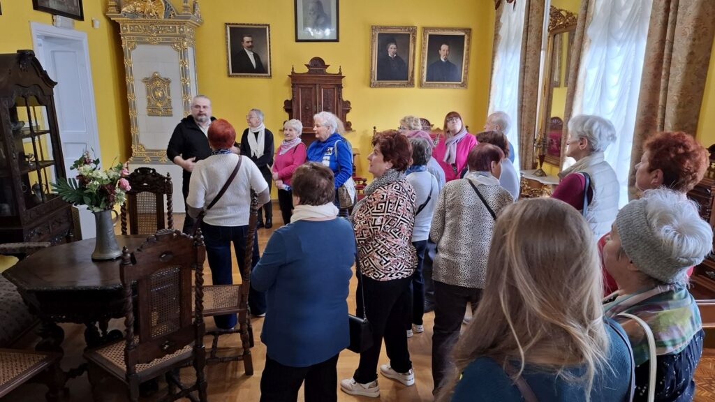 Grupa dorosłych uczestników zwiedzania słucha przewodnika w zabytkowym wnętrzu muzeum z portretami na ścianach (kliknięcie powiększy zdjęcie).