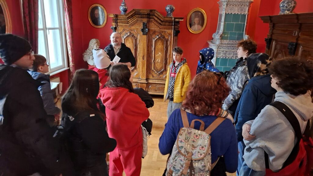 Przewodnik oprowadza grupę po sali z historycznymi meblami i eksponatami, uczestnicy stoją wokół i słuchają (kliknięcie powiększy zdjęcie).
