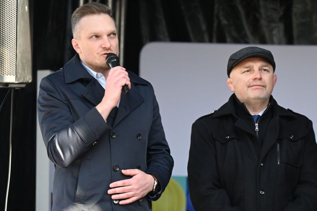 Mężczyzna w płaszczu przemawia do publiczności przez mikrofon, obok stoi drugi uczestnik wydarzenia (kliknięcie powiększy zdjęcie).