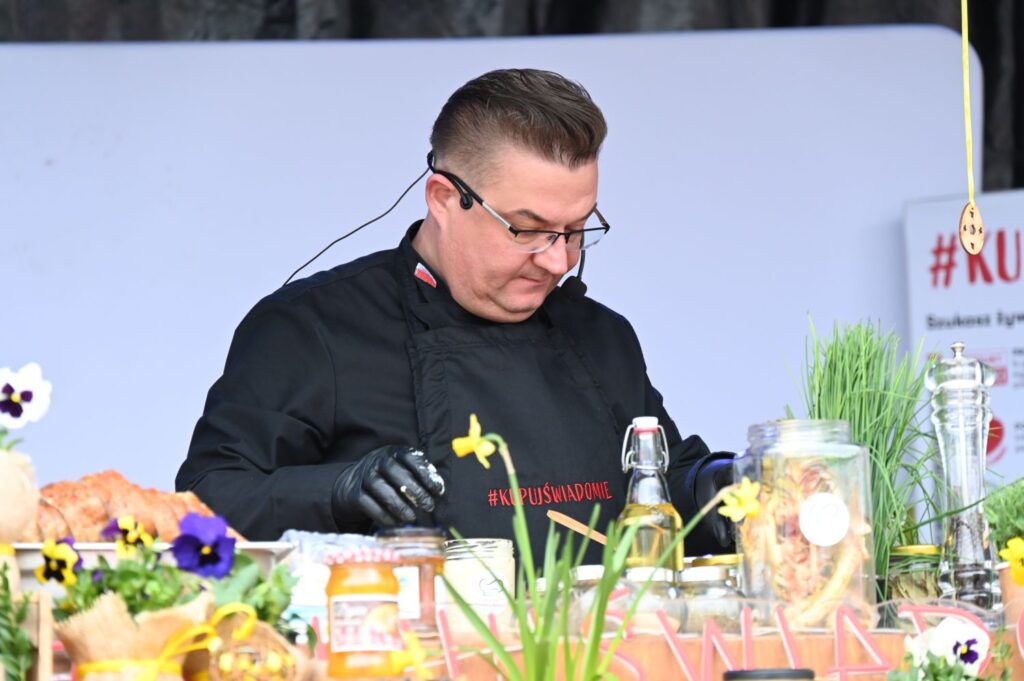 Kucharz przygotowuje składniki na stole pełnym produktów i wiosennych dekoracji podczas pokazu kulinarnego (kliknięcie powiększy zdjęcie).
