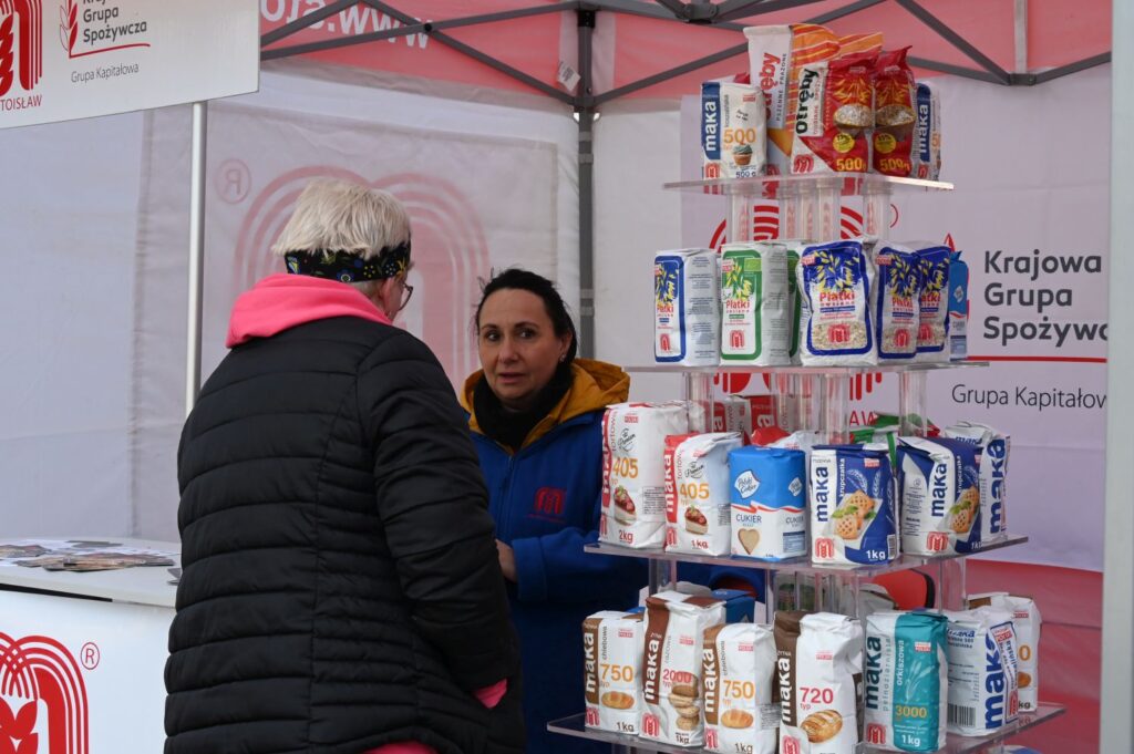 Stoisko z produktami spożywczymi, gdzie sprzedawczyni obsługuje klientkę podczas kiermaszu (kliknięcie powiększy zdjęcie).