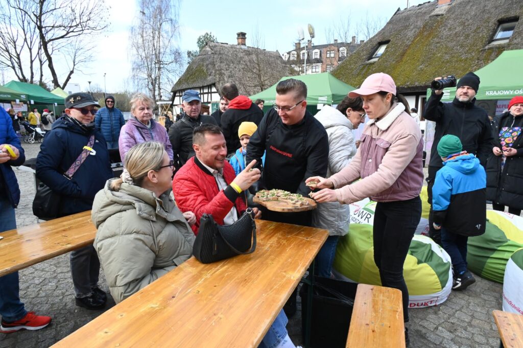 Uczestnicy siedzą przy stołach, a organizatorzy podają potrawy podczas wspólnej degustacji na kiermaszu (kliknięcie powiększy zdjęcie).