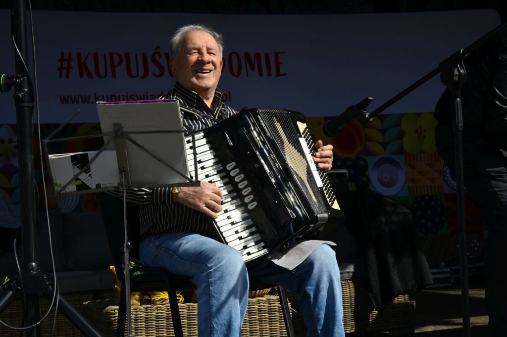 Starszy mężczyzna gra na akordeonie na scenie podczas kiermaszu, siedząc przy mikrofonie (kliknięcie powiększy zdjęcie).