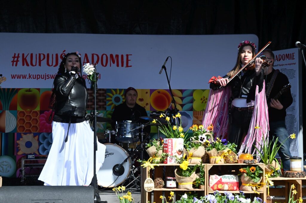 Wokalistka i skrzypaczka występują na scenie z zespołem w tle, przed nimi stoły z kwiatami, przetworami i ozdobami wielkanocnymi (kliknięcie powiększy zdjęcie).